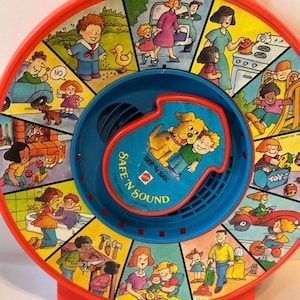 May include: Vintage red and blue Safe 'N Sound toy with a handle. The toy features a rotating wheel with colorful illustrations depicting various scenes. The center has a blue section with the text "Safe 'N Sound" and a picture of a boy and a dog.