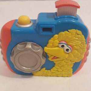 May include: A colorful toy camera featuring Big Bird from Sesame Street. The camera is primarily blue, with red and yellow accents. It has a red chimney-like structure on top and a large dial on the front.