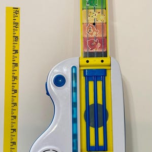 Pode incluir: Uma guitarra de brinquedo colorida com corpo branco e amarelo, detalhes azuis e um bot&atilde;o vermelho. A escala apresenta ilustra&ccedil;&otilde;es de animais. Uma r&eacute;gua amarela est&aacute; ao lado da guitarra. O brinquedo est&aacute; rotulado "baby einstein-".