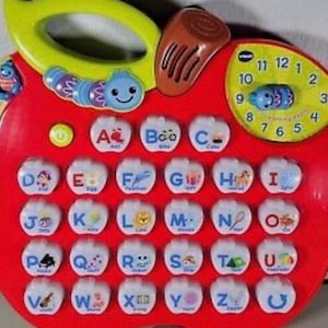 May include: A red, apple-shaped educational toy with a yellow handle and a clock face. It features alphabet letters and a caterpillar design. The toy is designed for early learning.