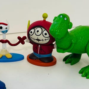Peut inclure: Une collection de figurines de personnages color&eacute;s. Les figurines comprennent une fourchette blanche avec un cure-pipe rouge et marron, une figurine &agrave; capuche rouge avec trois yeux et un jouet dinosaure vert. Les jouets sont sur une surface blanche.