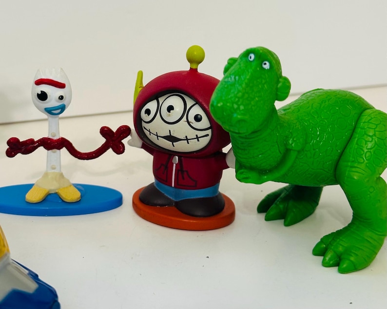 Peut inclure: Une collection de figurines de personnages color&eacute;s. Les figurines comprennent une fourchette blanche avec un cure-pipe rouge et marron, une figurine &agrave; capuche rouge avec trois yeux et un jouet dinosaure vert. Les jouets sont sur une surface blanche.