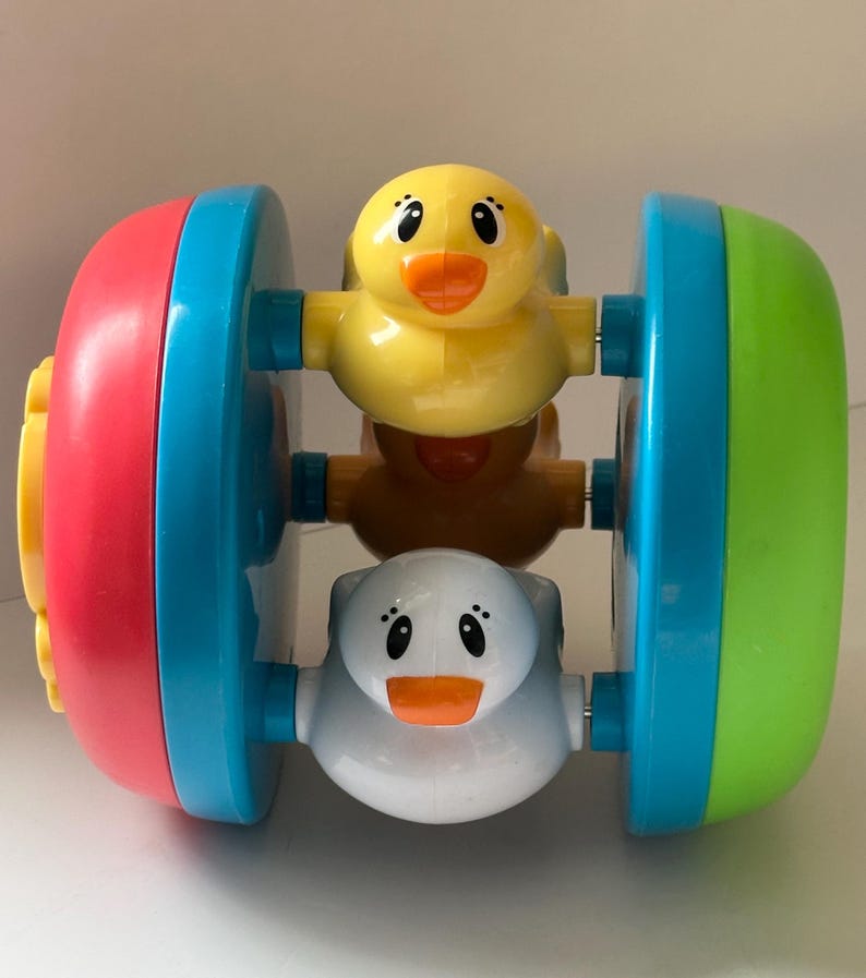 May include: A colorful baby toy featuring three duck figures. The toy has a cylindrical shape with red, blue, and green ends. The ducks are yellow, brown, and white with orange beaks. The toy is designed for infants and toddlers.