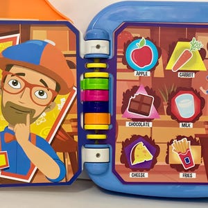 May include: A colorful children's toy book with a blue and orange frame. The left side features a cartoon character with the text "This is so much fun!" The right side displays food illustrations with labels like "Apple" and "Ice Cream."