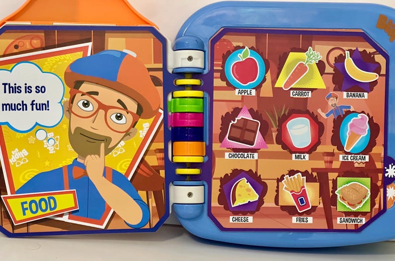 May include: A colorful children's toy book with a blue and orange frame. The left side features a cartoon character with the text "This is so much fun!" The right side displays food illustrations with labels like "Apple" and "Ice Cream."