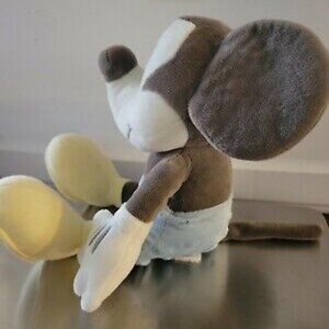 May include: A plush toy of a cartoon mouse, featuring a white face and hands, large gray ears, and a brown body. The mouse is wearing light blue shorts and has yellow feet. The toy is sitting down.