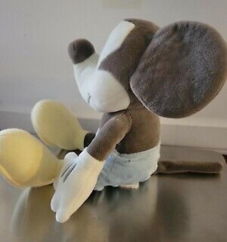 May include: A plush toy of a cartoon mouse, featuring a white face and hands, large gray ears, and a brown body. The mouse is wearing light blue shorts and has yellow feet. The toy is sitting down.