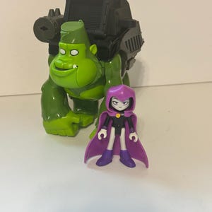 May include: A green gorilla action figure with a black mechanical backpack component positioned behind a small purple-cloaked figure with a black outfit, white skin, and a yellow circular emblem on the chest, standing on a plain surface.