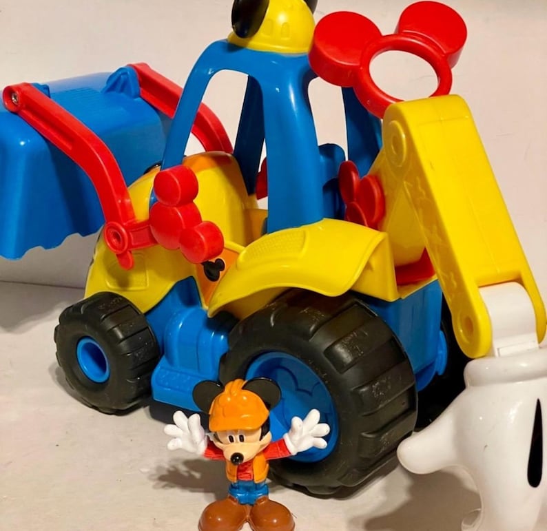 May include: A colorful toy construction vehicle with a Mickey Mouse figure. The vehicle is primarily blue, yellow, and red, with black tires. The Mickey Mouse figure is wearing an orange hard hat and work clothes. The toy is designed for children.