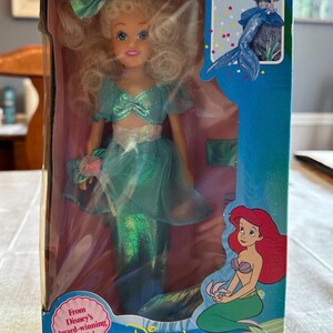 May include: A boxed doll of Ariel's sister Arista from Disney's The Little Mermaid. The doll has blonde hair and wears a teal outfit with a mermaid tail. The box features the movie's logo and the text "Ages 3 and up."