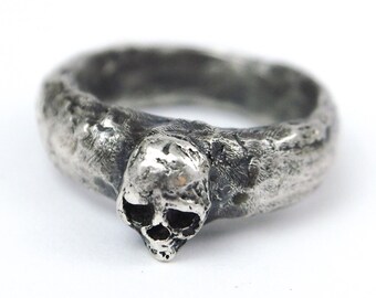 Può includere: Un anello d'argento a forma di teschio con una fascia testurizzata. Il teschio è dettagliato e ha un aspetto leggermente usurato.