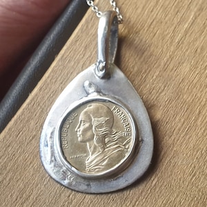 May include: A silver teardrop-shaped pendant with a gold coin inside. The coin features a profile of a woman with the text "REPUBLIQUE FRANCAISE" around the edge.