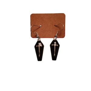 May include: A pair of black coffin-shaped earrings with a gold cross design. The earrings are hanging from a brown card.