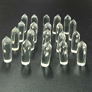 May include: A collection of eighteen clear, colorless, faceted crystal points arranged in a grid. Each individual piece features a pointed, multi-faceted top and a cylindrical body with vertical facets, reflecting light across their surfaces.