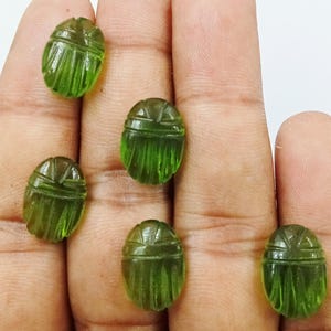 May include: Six translucent green scarab beetle gemstones. Each gemstone is oval-shaped with carved details, including a segmented body and head. The gemstones are displayed on a hand against a white background.