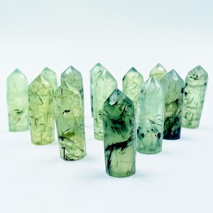 May include: A collection of green quartz crystal points with dark green inclusions. The crystals are arranged on a white surface, each with a pointed top and a cylindrical base. The crystals vary in size and the inclusions create a unique pattern in each.