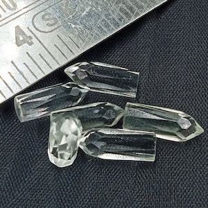 May include: Six clear quartz crystal points, also known as wands, are arranged on a dark gray fabric surface. The crystals are faceted and have a pointed tip.