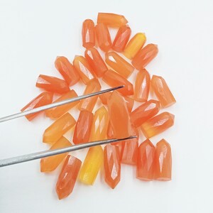 May include: A collection of faceted, orange-colored gemstone crystals. The crystals are in various shades of orange and yellow, with a pointed, elongated shape. A pair of silver tweezers is holding one of the crystals. The crystals are scattered on a white surface.
