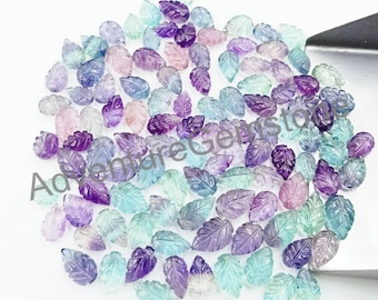 Puede incluir: Una colección de aproximadamente 100 piedras preciosas sueltas, talladas en forma de hoja, en tonos de morado, azul y verde. Las piedras preciosas están esparcidas sobre una superficie blanca.