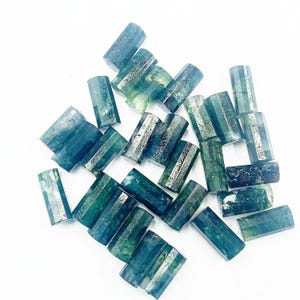 May include: A collection of small, cylindrical, polished gemstone beads in various shades of deep blue, teal, and forest green. Each bead features a smooth, hexagonal-cut surface with natural internal inclusions and variations in color intensity.