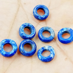 Könnte beinhalten: Sieben blaue und weiße Lapislazuli-Donut-Perlen. Die Perlen sind glatt und poliert mit einer hellblauen Farbe und weißen Marmorierungen.