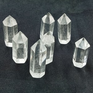 May include: Seven clear quartz crystal points, also known as clear quartz wands, are arranged on a black fabric background. The points are all approximately the same size and shape, and they are all pointed at the top.