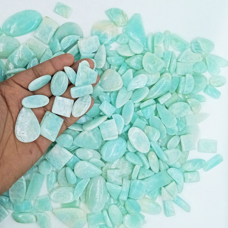 K&ouml;nnte beinhalten: Eine gro&szlig;e Sammlung von hellblauen und gr&uuml;nen Edelstein-Cabochons in verschiedenen Formen und Gr&ouml;&szlig;en. Die Cabochons sind auf einer wei&szlig;en Oberfl&auml;che zu einem Haufen angeordnet.