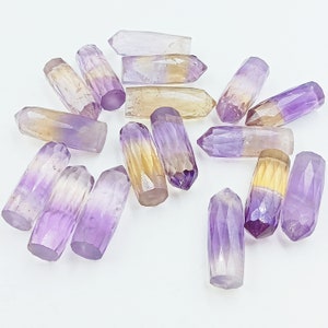 May include: A collection of faceted amethyst crystal points in various shades of purple, yellow, and clear. The crystals are cut into a cylindrical shape with a pointed tip. The crystals are arranged on a white surface.