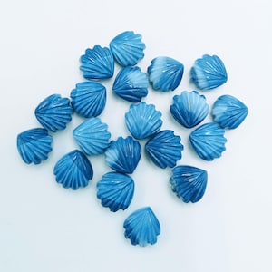 Puede incluir: Una colección de cuentas azules en forma de concha con un efecto veteado blanco. Las cuentas están dispuestas sobre una superficie blanca. Estas cuentas decorativas son ideales para la fabricación de joyas y proyectos de artesanía.