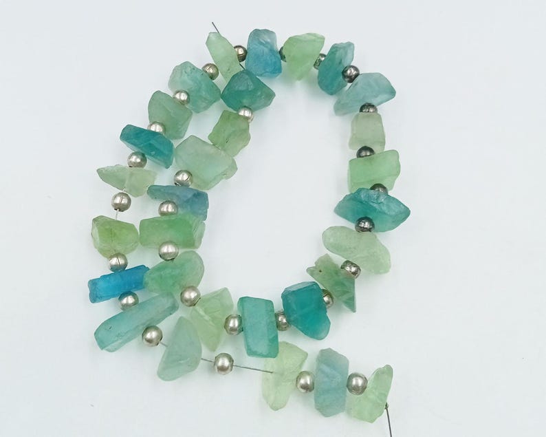 Op de afbeelding: Een streng teal en groene edelsteen kralen met zilveren spacer kralen. De kralen zijn onregelmatig van vorm en hebben een natuurlijke, ruwe textuur.