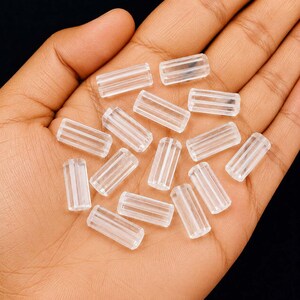 May include: A collection of small, clear, cylindrical beads with a ribbed, grooved texture along their length. These transparent, tube-shaped beads are scattered across the palm, showcasing their uniform size and reflective, glass-like appearance.
