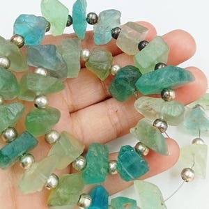 May include: A strand of green and blue gemstone beads with silver spacers. The beads are irregular in shape and have a natural, rough texture.