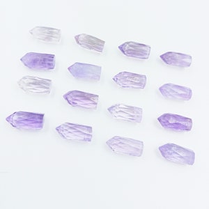 May include: A collection of 15 amethyst crystal points arranged in rows on a white background. The points are all different sizes and shapes, but they are all a light purple color.