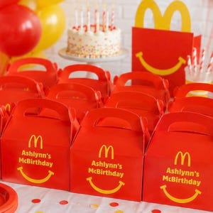 May include: Red McDonald's themed party favor boxes with handles, featuring the McDonald's logo, a yellow smiling face, and the text "Ashlynn's McBirthday". A birthday cake with candles and balloons are in the background.