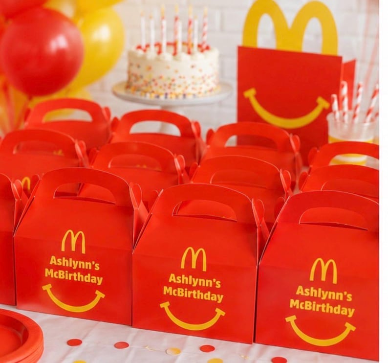 May include: Red McDonald's themed party favor boxes with handles, featuring the McDonald's logo, a yellow smiling face, and the text "Ashlynn's McBirthday". A birthday cake with candles and balloons are in the background.