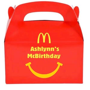 May include: A red cardboard party favor box with a handle. The box features the McDonald's golden arches logo, the text "Ashlynn's McBirthday", and a yellow smiling face. The box is designed for a birthday celebration.