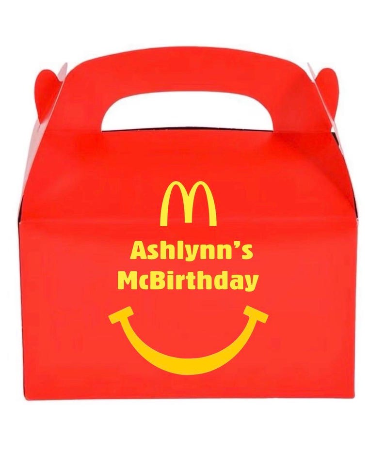 May include: A red cardboard party favor box with a handle. The box features the McDonald's golden arches logo, the text "Ashlynn's McBirthday", and a yellow smiling face. The box is designed for a birthday celebration.