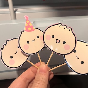 May include: Four dumpling-shaped cupcake toppers with smiling faces and pink cheeks. One topper has a pink and yellow party hat. The toppers are attached to wooden sticks, perfect for decorating cupcakes or other baked goods.