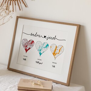 May include: A framed print featuring three heart-shaped map designs in red, teal, and yellow, with dates and text below each. The print is in a light wood frame, with the names "sandra & jacob" written above the hearts.