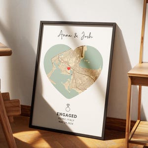 May include: Framed print featuring a map of Rome, Italy, shaped like a heart. The map is in shades of brown and beige, with a red heart marking a location. The text "Anna & Josh" is at the top, and "Engaged" with the date May 29, 2024, is at the bottom.