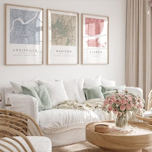 Puede incluir: Tres impresiones enmarcadas de mapas de ciudades cuelgan sobre un sofá blanco con cojines decorativos. Los mapas son de Louisville, Madison y Lisboa, cada uno con un esquema de color diferente. Una mesa de centro redonda de madera y una silla de mimbre completan la sala de estar.