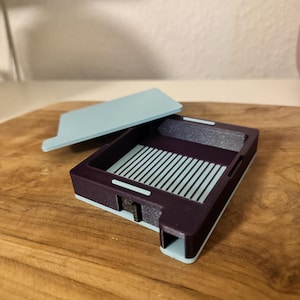 Könnte beinhalten: Eine kleine, rechteckige Box mit einem hellblauen Deckel. Die Box ist aus dunkelviolettem Kunststoff und hat ein Gittermuster im Inneren. Der Deckel ist leicht geöffnet und gibt den Blick auf das Innere der Box frei.