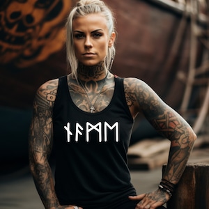 Può includere: Una donna con tatuaggi indossa una canotta nera con la scritta "FAMM" in bianco sul davanti. La canotta ha un collo rotondo e spalline sottili.