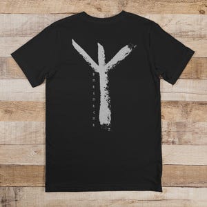 Può includere: T-shirt nera con un grande simbolo runico grigio consumato. Sotto la runa, le lettere "B M R E M R M R" sono allineate verticalmente. La maglietta è esposta su una superficie di legno.