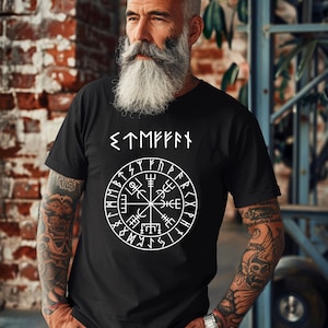 May include: Black t-shirt with a white Viking compass design and runes. The shirt features a circular symbol with various lines and shapes, and text above the symbol. The shirt is made of a soft material.