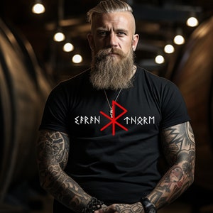 Può includere: Una maglietta nera con un simbolo rosso e il testo "EFRN TNORM" stampato sul davanti. La persona che indossa la maglietta ha una lunga barba e tatuaggi sulle braccia.