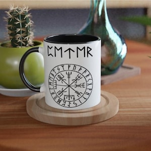 Könnte beinhalten: Weiße Keramiktasse mit schwarzem Rand und Henkel. Die Tasse zeigt ein schwarz-weißes Design eines Kompasses mit Runen um den Außenrand. Der Text "CMTMR" ist oben auf der Tasse aufgedruckt.