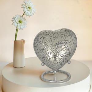 May include: A silver heart-shaped cremation urn with a floral design. The urn is on a silver stand.