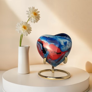 May include: A heart-shaped ceramic urn with a swirling blue, red, and white design. The urn is on a gold metal stand.