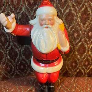 Puede incluir: Una figura vintage de Papá Noel, con un traje rojo, barba blanca y un gorro rojo con un emblema dorado. La figura tiene un gesto de bienvenida con un brazo extendido. Decoración navideña clásica.